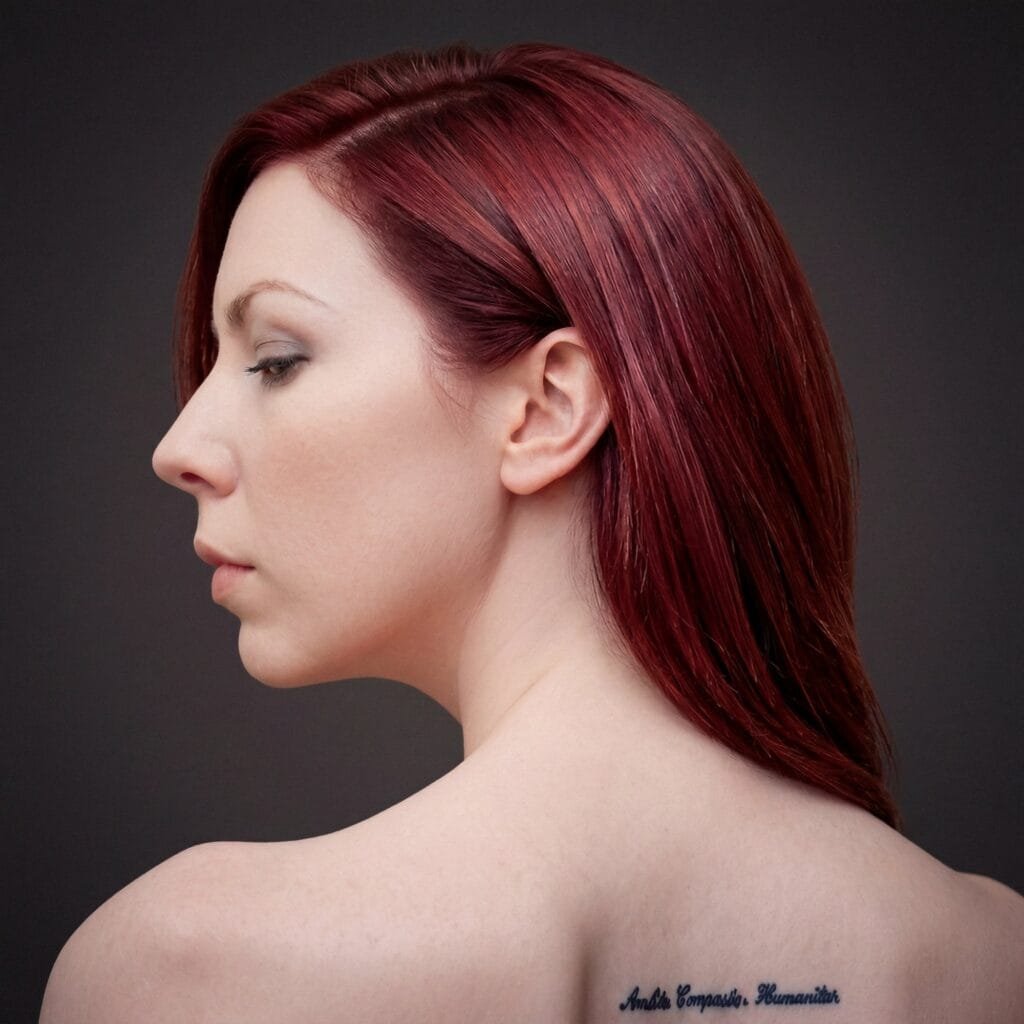 A studio portrait of a woman with deep red hair, turned away from the camera with her face brought back round in profile, bare shoulders, against a dark grey ground in soft cool light. A line of small Latin script runs across her shoulder blade.