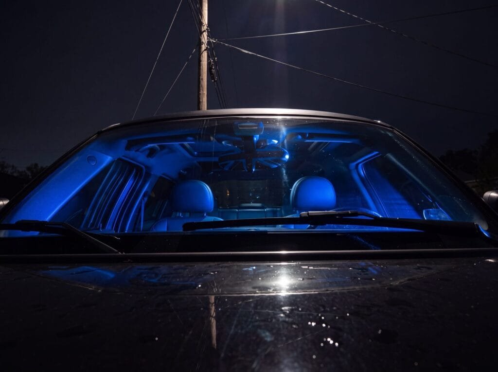 A MINI Cooper photographed head-on through its windshield on a wet night, the cabin glowing electric blue from photographic panels laid on the seats, a utility pole and wires rising behind the roof