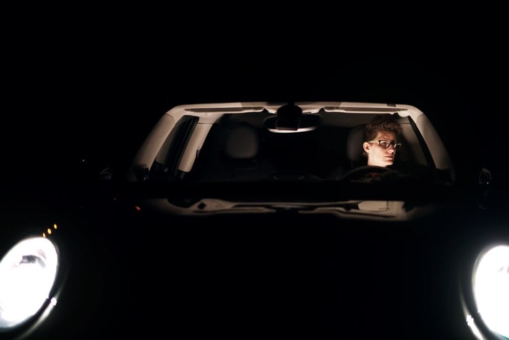 The same MINI Cooper almost entirely in darkness, only its two round headlights and a faintly lit cabin visible, with the owner at the wheel